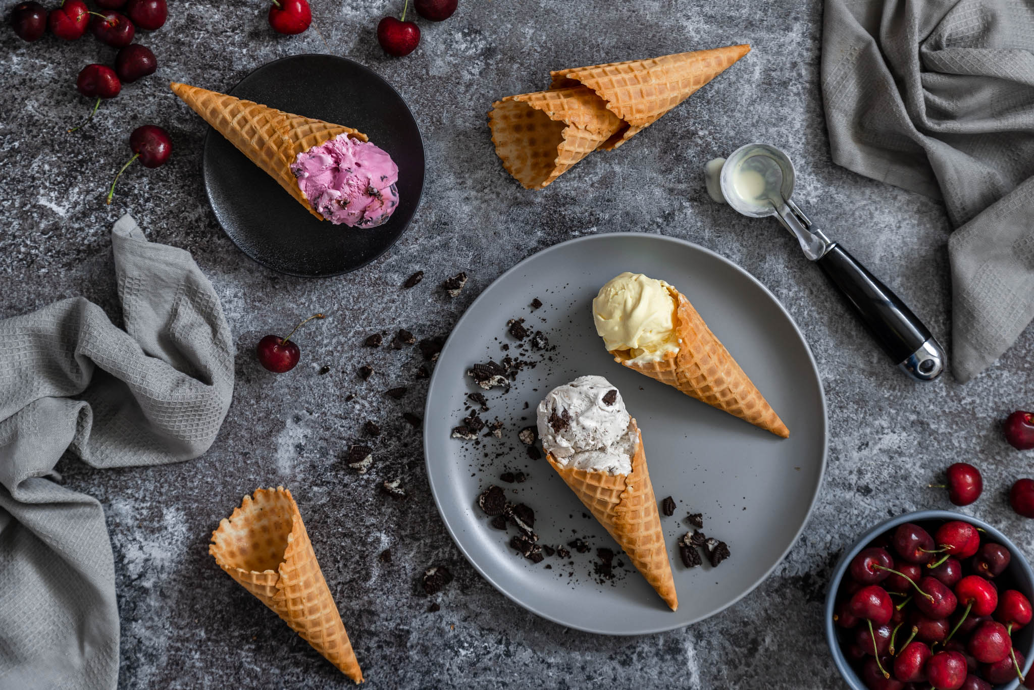 Flat lay of Chapman's Ice Cream in sugar cones along with ice cream spoon and black cherries.